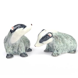 May include: A pair of ceramic badger figurines, one looking up and the other forward. The badgers have black and white striped heads, gray fur, and small pink ears. These decorative figurines are ideal for a home decor collection.