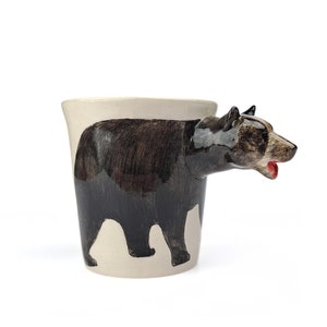 May include: A ceramic mug with a black bear design. The bear's head is the handle of the mug. The bear is painted in shades of brown and black.
