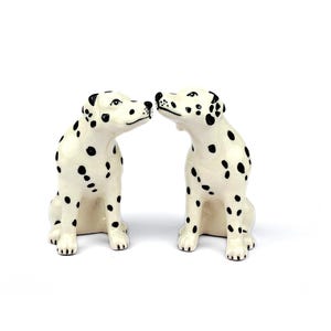 May include: Two ceramic Dalmatian dog ornaments. The dogs are white with black spots and are facing each other. The ornaments are sitting upright with their noses almost touching. The background is plain white.