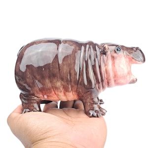 May include: A ceramic figurine of a brown and pink hippopotamus with a wide open mouth. The hippopotamus is standing on a person's hand.