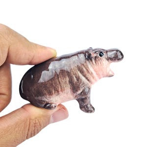 May include: A small, ceramic hippo figurine in shades of brown and pink. The hippo has its mouth open, revealing a pink interior. The figurine is held in a hand against a white background.