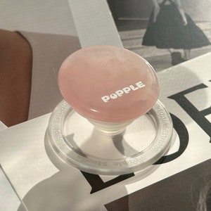 May include: A pink PopSockets phone grip with a white base and a clear ring. The grip is made of a smooth, polished material and has the word "PopSockets" printed on it.