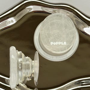 Könnte beinhalten: Zwei transparente Handygriffe aus Kunststoff, einer ist ein PopSocket-Markengriff mit dem Wort "PopSocket" darauf gedruckt, der andere ist ein transparenter Griff mit einem Stapel Ringe.