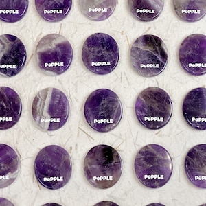 Könnte beinhalten: Vierundzwanzig runde, polierte Amethyststeine, jeder mit einem einzigartigen Muster aus Lila und Weiß. Die Steine sind in vier Reihen zu sechs angeordnet, wobei das Wort "POPPLE" auf jedem Stein gedruckt ist.