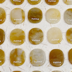 Könnte beinhalten: Eine Sammlung von 20 glatten, ovalen Steinen in Gelbbraun. Jeder Stein trägt die Aufschrift "POPPLE" in Weiß.