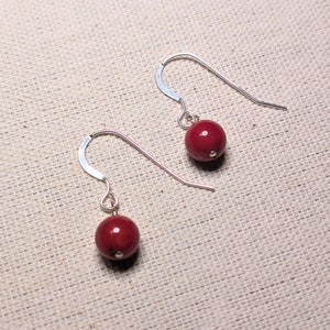 May include: A pair of drop earrings. Each earring has a deep red, spherical bead hanging from a silver-coloured hook. The beads have a smooth, glossy finish. The earrings are set against a neutral, textured background.