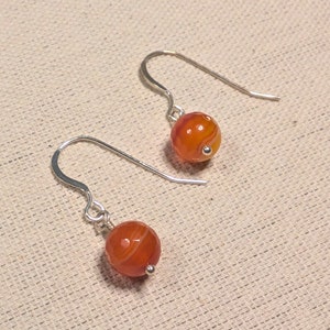 May include: A pair of earrings featuring orange and white striped beads. The beads are round and dangle from silver-colored ear wires. The earrings are displayed on a neutral-colored fabric background.
