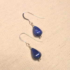 Puede incluir: Un par de pendientes con piedras de lapislázuli azul oscuro en forma de lágrima. Las piedras cuelgan de ganchos plateados. Los pendientes se muestran sobre una superficie neutra y texturizada.