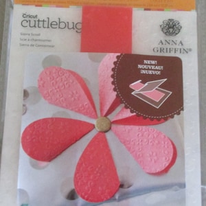May include: A pink and white flower-shaped die cut for use with a Cricut Cuttlebug die-cutting machine. The die cut is labeled "New! Nouveau! Inuevo!" and is a product of Anna Griffin.