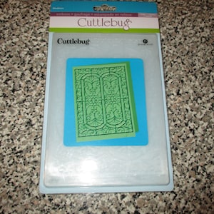 May include: A white plastic package with a blue border contains a green and white die-cutting template for a decorative design. The package is labeled "Cuttlebug" and "Made in USA".