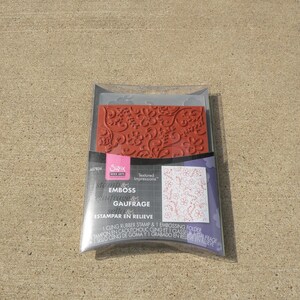 May include: A clear plastic package containing a red rubber stamp and a clear plastic embossing folder. The stamp features a floral design with swirls and flowers. The text on the package reads "Sizix Textured Impressions stamp & emboss gaufrage sello estampar en relieve 1 cling rubber stamp & 1 embossing folder tampon en caoutchouc cling et 1 classeur a gaufrage sello cling de goma y 1 grabado en relieve".