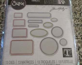 May include: A sheet of 13 metal dies for crafting, featuring various oval and rectangular shapes with decorative borders. The dies are designed by Jim Holtz and are part of the Sizzix Thinlits collection. The text "13 DIES | 13 MATRICES | 13 TROQUELES | 13 FUSTELLE | 13 ВЫРУБКИ | 13 STANZEN | 13 CORTANTES" is printed at the bottom.