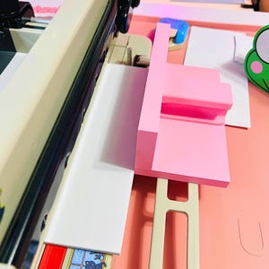 May include: A beige paper cutting machine featuring a long metal blade and a sliding mechanism. A bright pink plastic guide block is positioned on the cutting surface to align white paper sheets. The machine has a sturdy base and a handle.