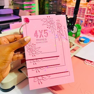 May include: A set of four stacked pink rectangular acrylic templates held together by a metal ring. Each template features a floral border design and the text 4X5 INCHES 101MMX127MM printed in a glittery finish. The surface has a subtle holographic sparkle.