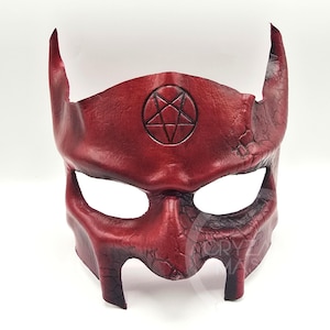 May include: A deep red leather mask featuring two prominent upward-curving horns and a central embossed pentagram symbol. The surface displays a textured, cracked finish with darker shading, and the design includes two large eye openings.