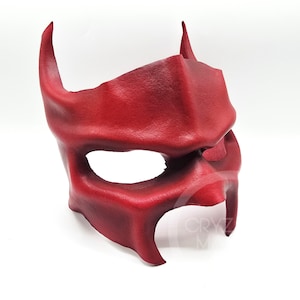 May include: A deep red moulded mask featuring a textured surface, pointed ear-like projections on the sides, a prominent central ridge, and wide eye openings. The mask has a structured, angular design with a watermark reading CRYZ M.