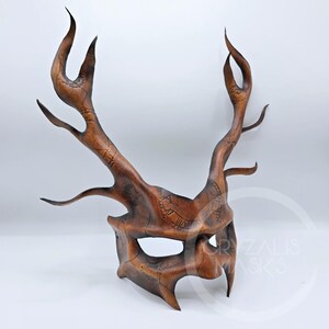 May include: A brown leather mask featuring tall, branching antler-like protrusions extending upwards. The surface is etched with intricate clockwork patterns and Roman numerals. The text CRYZALIS MASKS is visible in the lower right corner.