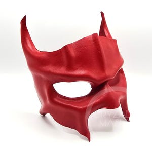 May include: A handcrafted, red leather mask with a devil-like design. The mask features two curved horns and a pointed top, with eye openings and a contoured shape for the face. The mask is made of a smooth, solid material.