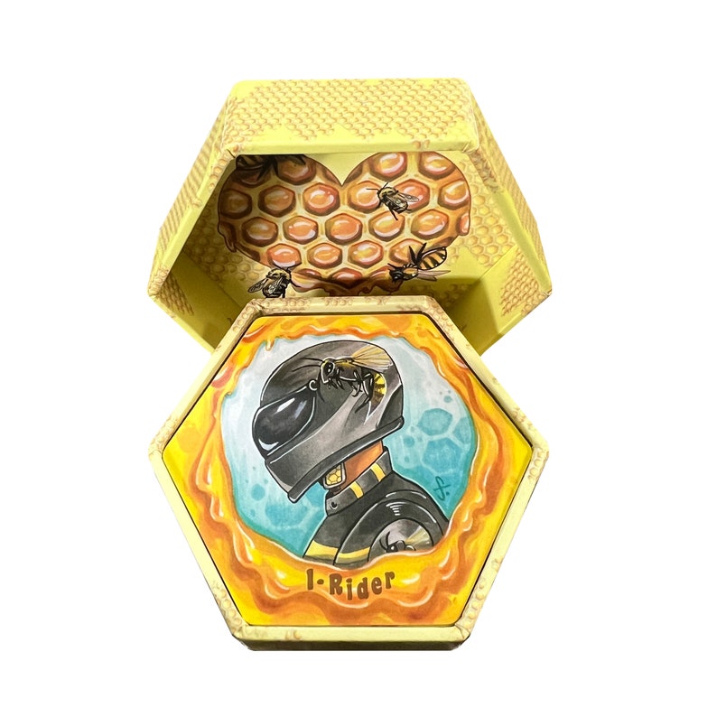 May include: A hexagonal box with a yellow honeycomb pattern. The box is open and reveals a yellow honeycomb heart with bees. The bottom of the box has a drawing of a person wearing a black helmet and a black and yellow suit. The text "1-Rider" is printed on the bottom of the box.