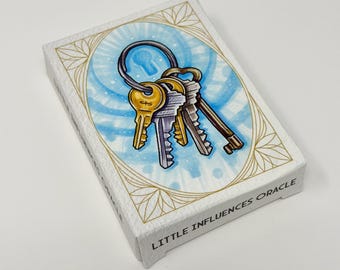 May include: A deck of cards titled "Little Influences Oracle" features an illustration of keys on a ring. The keys are gold, silver, and bronze, set against a blue and white background with a keyhole design. The deck has a white border with gold accents.