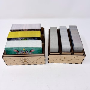 May include: Two light-colored wooden storage boxes for card decks. The left box features an engraved eye and star design, holding three stacks of cards. The right box features an engraved crescent moon and star design, holding three stacks of cards.