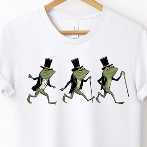Könnte beinhalten: Weißes T-Shirt mit einer skurrilen Illustration von drei grünen Fröschen mit Zylinder, Smoking und Spazierstock. Die Frösche sind in Bewegung, was auf ein verspieltes, animiertes Design hindeutet. Das Shirt wird an einem Holzbügel präsentiert.