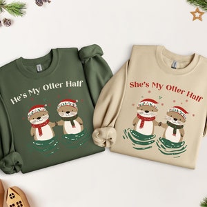Könnte beinhalten: Zwei grüne und beige Weihnachtspullover mit einem passenden Design aus zwei Ottern, die Weihnachtsmützen tragen und sich an den Händen halten. Der Text auf dem grünen Pullover lautet "He's My Otter Half" und der Text auf dem beigefarbenen Pullover lautet "She's My Otter Half".