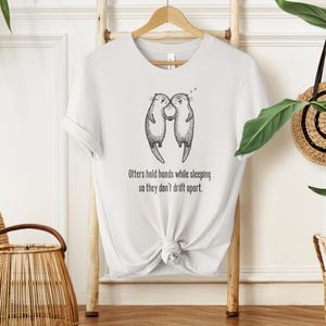 Peut inclure: T-shirt blanc avec un dessin de deux loutres se tenant la main, avec le texte "Otters hold hands while sleeping so they don't drift apart." Le t-shirt est suspendu à un cintre en bois.