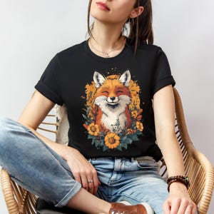 una donna seduta con una maglia nera con l'immagine di una volpe sorridente circondata da fiori in stile cottagecore