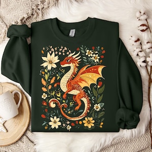 Peut inclure: Un sweat-shirt vert foncé avec un motif floral mettant en scène un dragon rouge et orange. Le dragon est entouré de fleurs et de feuilles dans des tons de jaune, orange et vert.