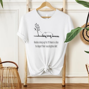 Può includere: T-shirt bianca con un'illustrazione nera di un koala che dorme sotto un ramo d'albero. Il testo recita: "I koala dormono fino a 20 ore al giorno per digerire la loro dieta di eucalipto."