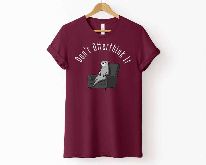 una tshirt color marroon con una lontra su una poltrona e sopra di lei la scritta "Don't otterthink it"