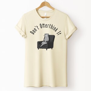 una tshirt color naturale con una lontra su una poltrona e sopra di lei la scritta "Don't otterthink it"