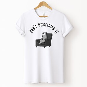 una tshirt bianca con una lontra su una poltrona e sopra di lei la scritta "Don't otterthink it"