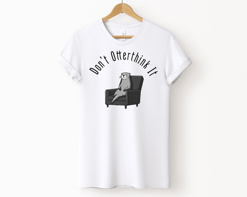 una tshirt bianca con una lontra su una poltrona e sopra di lei la scritta "Don't otterthink it"
