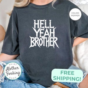 May include: Pepper-colored t-shirt with the text "HELL YEAH BROTHER" in a white, stylized font. The shirt has a relaxed fit and short sleeves. The image also includes a small round tag that says "COLOR: PEPPER".