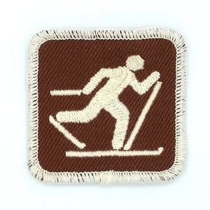 May include: A brown square patch with a white embroidered image of a person cross-country skiing. The patch has a white stitched border.