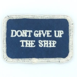 Puede incluir: Un parche de tela azul con costuras blancas alrededor de los bordes. El parche tiene texto blanco que dice "Don't Give Up The Ship".