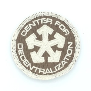 May include: A brown embroidered patch with white stitching. The patch is circular and features a white starburst with six points and the text "CENTER FOR DECENTRALIZATION" around the starburst.