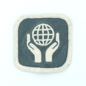 May include: A gray fabric patch with white stitching around the edges. The patch features a white outline of two hands holding a globe.