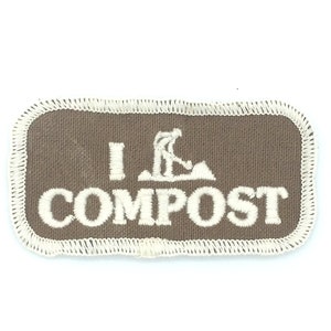 Può includere: Un cerotto in tessuto marrone con cuciture bianche attorno ai bordi. Il cerotto ha una silhouette bianca di una persona che scava in un cumulo di terra e il testo "I COMPOST" in bianco.