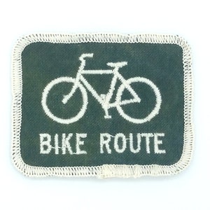 Könnte beinhalten: Grüner Stoff-Patch mit weißen Nähten und einem weißen Fahrrad-Symbol. Der Text "BIKE ROUTE" befindet sich unter dem Fahrrad.