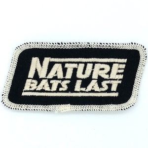 Puede incluir: Parche bordado en blanco y negro con el texto "NATURE BATS LAST" en blanco sobre fondo negro.