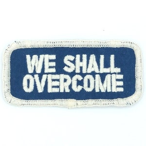 Peut inclure: Un patch rectangulaire bleu avec des coutures blanches autour des bords. Le patch dit "WE SHALL OVERCOME" en lettres blanches.