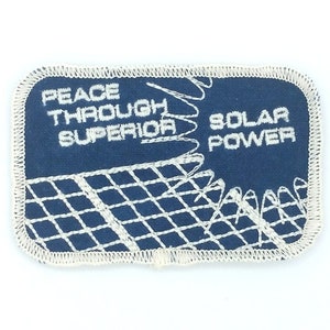Puede incluir: Un parche bordado azul con costuras blancas. El parche presenta un diseño de panel solar y el texto "Peace Through Superior Solar Power".