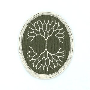 May include: An embroidered patch featuring a white tree design on a dark olive green background. The tree is centered within an oval shape, with a cream-colored border. The patch is likely for clothing or accessories.