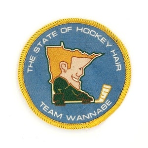 Może przedstawiać: Naszywka haftowana w kolorze niebieskim i żółtym z tekstem "The State of Hockey Hair Team Wannabe" i kreskówkowym obrazkiem hokeisty w zielonej koszulce trzymającego kij hokejowy.