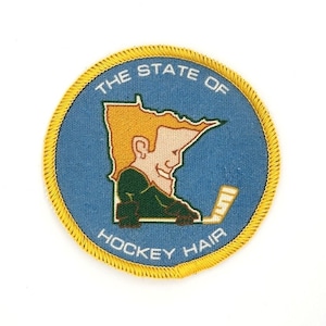 以下が含まれることがあります： 「The State of Hockey Hair」という文字と、ブロンドの髪をした人が緑のホッケーユニフォームを着てホッケーのスティックを持っているという漫画の画像が描かれた、青と黄色の刺繍パッチです。