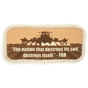 Op de afbeelding: Een rechthoekige, beige patch met een bruin silhouet van een tractor die een veld ploegt. De tekst "The nation that destroys its soil destroys itself." - FDR is gedrukt onder de tractor.