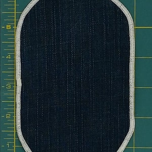 May include: Oval-shaped patch with a dark blue, vertically striped fabric center. The patch is outlined with a white, stitched border. The background is a green cutting mat with measurement markings.